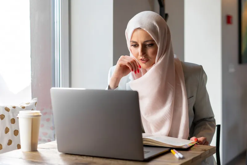 A woman in a pale pink hijab and grey blazer sits at her laptop, fingers at her chin, reading the screen beside an open notebook.
