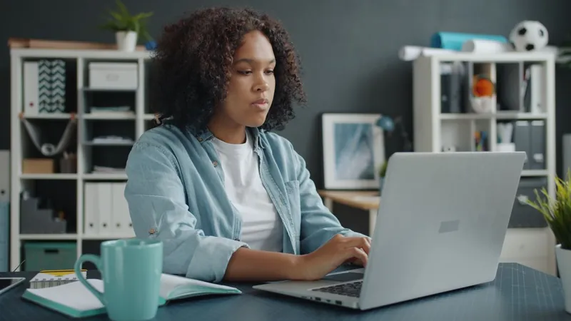 A woman with curly hair in a denim shirt types on her laptop at a desk, with white shelves behind her.