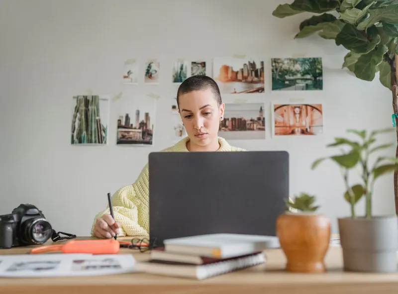 A woman works at her laptop with a pen in hand, her photographs pinned on the wall behind her.