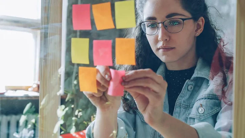 A woman in glasses placing a blank sticky note onto a glass planning wall covered in a grid of coloured notes.
