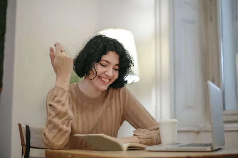 A woman with short dark curly hair smiles as she writes at a table beside her laptop.