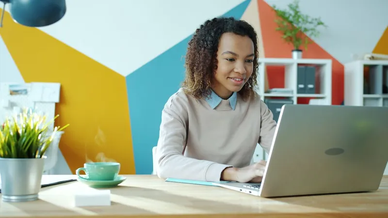 A woman with curly hair smiles at her laptop screen as she types at a desk, in an office with a colorful painted wall.